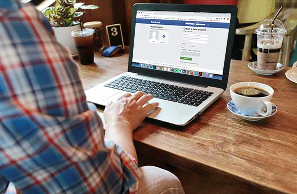 Man on the Facebook login page on his laptop, on a wooden table with a coffee next to the laptop